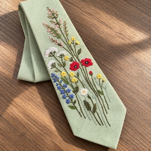 May include: A sage green necktie featuring detailed embroidery of wildflowers including red poppies, yellow blossoms, white clusters, and blue petals. The floral design extends vertically along the fabric, showcasing green stems and leaves.