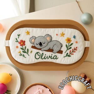 May include: A white fabric sleep mask featuring an embroidered gray koala sleeping on a brown branch, surrounded by colorful flowers and stars. The name Olivia is embroidered in green cursive script below the koala on the mask.