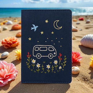 Puede incluir: Un portapasaporte azul marino con un dise&ntilde;o bordado de una furgoneta camper, luna, estrellas y flores. Un peque&ntilde;o p&aacute;jaro tambi&eacute;n est&aacute; bordado en la cubierta. El portapasaporte est&aacute; en una playa de arena con flores y conchas.