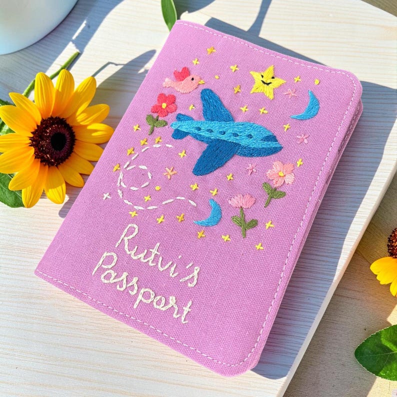 Puede incluir: Una funda de pasaporte lila con detalles bordados. Un avi&oacute;n azul, estrellas, flores y un p&aacute;jaro est&aacute;n bordados en la parte delantera. El texto "Rutvi's Passport" est&aacute; bordado en blanco. La funda est&aacute; cerrada y descansa sobre una superficie de madera.