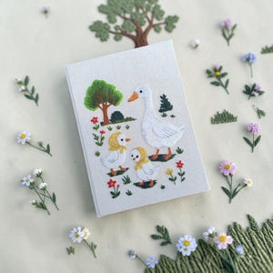 Puede incluir: Un cuaderno rectangular con cubierta de tela gris claro que presenta una escena bordada de una oca blanca grande y dos gansitos con capotas amarillas. El dise&ntilde;o incluye un &aacute;rbol verde, peque&ntilde;as flores rojas y hierba verde.
