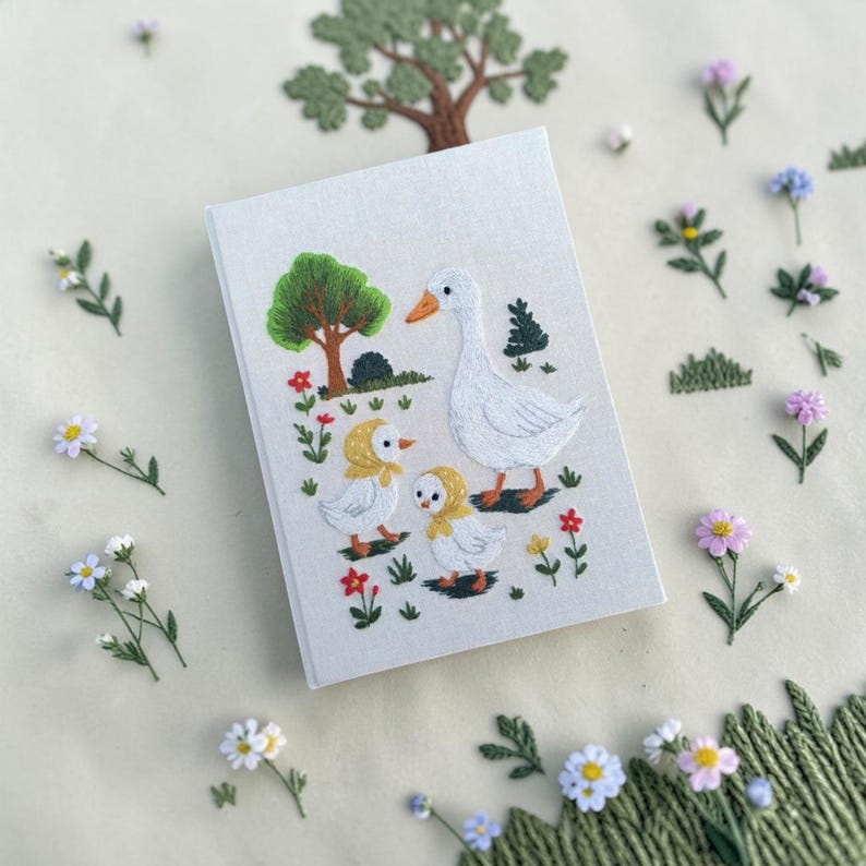 Puede incluir: Un cuaderno rectangular con cubierta de tela gris claro que presenta una escena bordada de una oca blanca grande y dos gansitos con capotas amarillas. El dise&ntilde;o incluye un &aacute;rbol verde, peque&ntilde;as flores rojas y hierba verde.