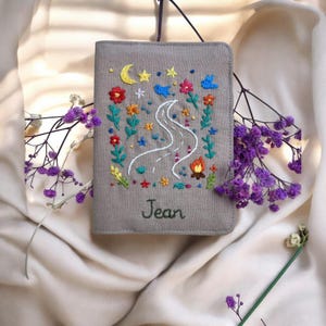 Puede incluir: Funda de pasaporte gris bordada con un dise&ntilde;o de carretera, fogata y flores. La cubierta presenta una luna creciente, estrellas y el nombre "Jean" bordado en cursiva. El soporte est&aacute; hecho de una tela suave y texturizada.