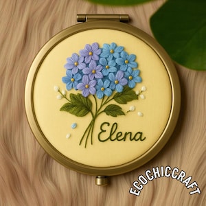 Puede incluir: Un espejo compacto redondo y dorado con un diseño floral bordado. El bordado muestra un ramo de flores azules y moradas con hojas verdes, el nombre "Elena" y el texto "ECOCHIC CRAFT".