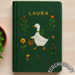 Puede incluir: Un cuaderno de tapa dura verde oscuro con detalles bordados de un ganso blanco que lleva una cesta marr&oacute;n, rodeado de flores naranjas y amarillas. El nombre LAURA est&aacute; bordado en amarillo en la parte superior y el texto ECOCHICCRAFT en la base.