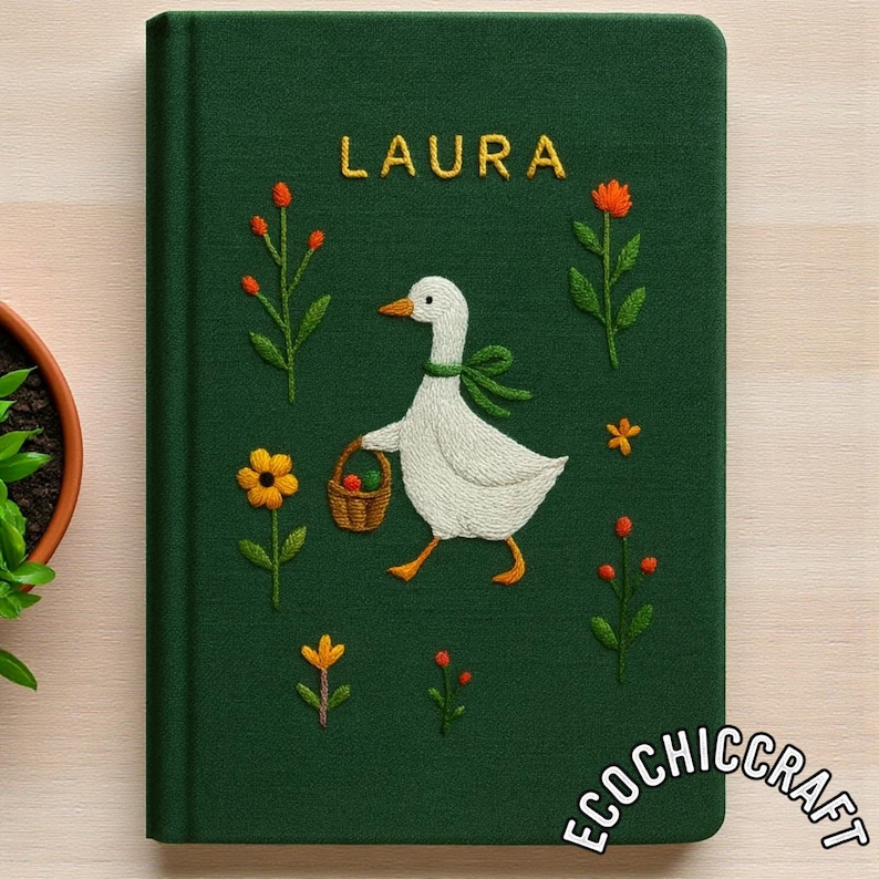 Puede incluir: Un cuaderno de tapa dura verde oscuro con detalles bordados de un ganso blanco que lleva una cesta marr&oacute;n, rodeado de flores naranjas y amarillas. El nombre LAURA est&aacute; bordado en amarillo en la parte superior y el texto ECOCHICCRAFT en la base.