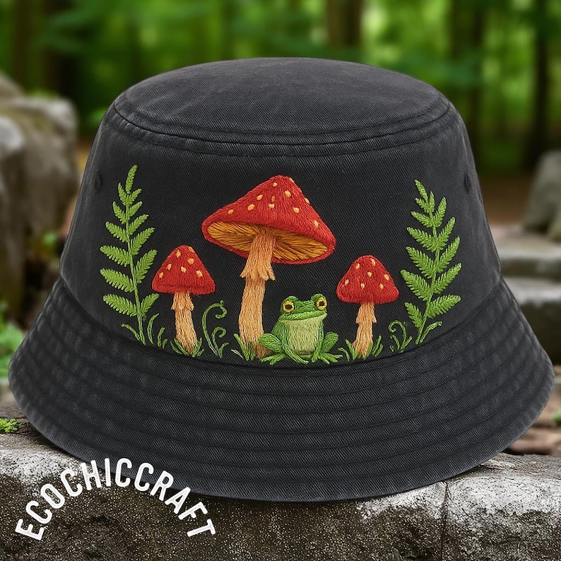 May include: A charcoal gray bucket hat featuring detailed embroidery of three red-capped mushrooms, a small green frog, and two green fern fronds. The text ECOCHICCRAFT is printed in white in the lower left corner of the image.