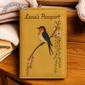May include: Mustard yellow passport cover with embroidered bird and floral design. The cover reads "Lana's Passport" in brown script. The bird has orange, black, and white details. The cover is rectangular and has a stitched border.