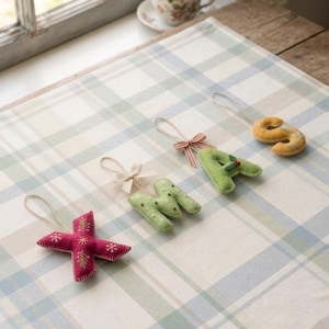Pode incluir: Quatro ornamentos de feltro em forma de letras que formam a palavra XMAS, cada um com um laço para pendurar. O X é rosa escuro com bordados dourados, o M é verde claro com pontos brancos, o A é verde claro com azevinho e o S é amarelo mostarda.