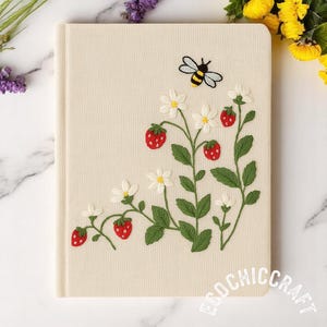 以下が含まれることがあります： 刺繍されたイチゴ、白い花、緑の茎、そして蜂が描かれたクリーム色のジャーナル。ジャーナルの表紙には、右下に円形のデザインで「ECOCHICCRAFT」というテキストが描かれています。