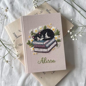 May include: A light beige fabric-covered notebook featuring an embroidered design of a black and white cat sleeping on a stack of books surrounded by white flowers and green leaves. The name Alissa is embroidered in green script at the bottom.