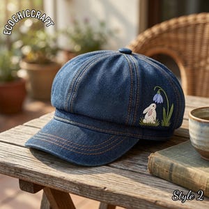 May include: A dark blue denim newsboy cap featuring an embroidered design of a white rabbit sitting next to a blue flower on the side. The cap has orange contrast stitching and the text ECOCHICCRAFT is visible in the upper left corner.