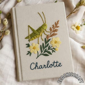 May include: A beige journal with an embroidered design featuring a green grasshopper, yellow flowers, and brown foliage. The name "Charlotte" is embroidered in blue script. The journal is a handmade item.