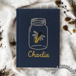Puede incluir: Un diario azul marino con un diseño bordado de una luciérnaga dentro de un tarro. El nombre "Charlie" está bordado en amarillo debajo del tarro. El diario está sobre una tela blanca.