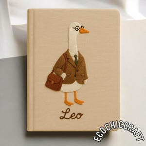 Puede incluir: Un cuaderno beige con una oca blanca bordada que lleva un blazer marr&oacute;n, una corbata verde y gafas redondas, cargando un malet&iacute;n marr&oacute;n. El nombre Leo est&aacute; bordado en letra cursiva marr&oacute;n debajo de la oca, con el texto ECOCHICCRAFT en la esquina.