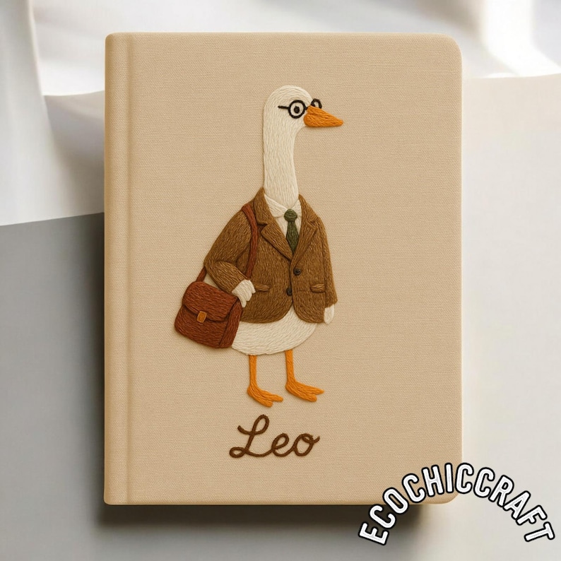 Puede incluir: Un cuaderno beige con una oca blanca bordada que lleva un blazer marr&oacute;n, una corbata verde y gafas redondas, cargando un malet&iacute;n marr&oacute;n. El nombre Leo est&aacute; bordado en letra cursiva marr&oacute;n debajo de la oca, con el texto ECOCHICCRAFT en la esquina.
