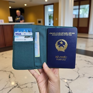 Puede incluir: Cartera de pasaporte verde azulado con un pasaporte vietnamita azul y tarjetas de cr&eacute;dito. El pasaporte tiene letras doradas y un emblema dorado. El soporte tiene bolsillos para tarjetas y divisas. El texto del pasaporte dice "PASSPORT".