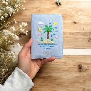 Puede incluir: Un portapasaporte azul claro con un bordado de una escena de isla tropical. El dise&ntilde;o incluye una palmera, luna, estrellas y flores. La palabra "Gree" est&aacute; bordada en la parte inferior. El portapasaporte est&aacute; en una mano.