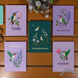 May include: A collection of notebooks with embroidered hummingbird designs. The notebooks are in various colors, including purple and green. Each notebook features a different hummingbird illustration and text, such as "Violet" and "Hummingbird".