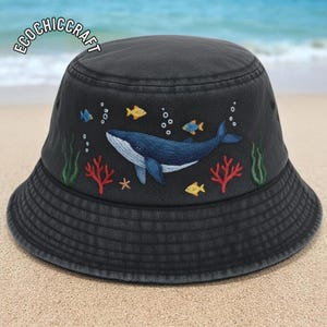 May include: Black bucket hat featuring an embroidered underwater design. A blue whale swims with yellow and blue fish, red coral, and green seaweed. The hat has the text "ECOCHIC CRAFT" on the top left.