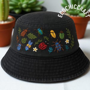 Op de afbeelding: Zwarte vissershoed met geborduurde kleurrijke insecten, bladeren en bloemen. De hoed heeft een brede rand en de tekst "ECOCHICCRAFT" in het wit. De borduursels bevatten insecten in blauw, geel, rood en groen.