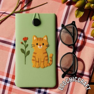 Könnte beinhalten: Eine hellgrüne Handyhülle mit einem orangefarbenen Tabby-Katzen-Design und einer roten Blume. Die Hülle hat einen schwarzen Knopfverschluss und wird mit einer Brille mit schwarzem Rahmen kombiniert. Der Text "ECOCHIC CRAFT" ist sichtbar.