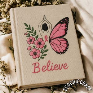 Könnte beinhalten: Ein beiges Notizbuch mit einem gestickten Design mit einem rosa Schmetterling, Blumen und dem Wort "Believe". Das Design beinhaltet eine Figur in einer Yoga-Pose und ein rosa Band. Das Notizbuch ist ein Buch mit Hardcover.