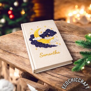 May include: A cream-coloured journal featuring an embroidered design of a yellow crescent moon, dark blue clouds, and stars. The name "Samantha" is embroidered in gold. The journal rests on a wooden surface.