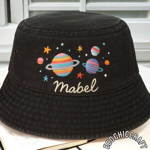Puede incluir: Sombrero de pescador negro con bordados de planetas azules, naranjas y amarillos. La palabra "Mabel" está bordada en cursiva. El sombrero tiene un diseño casual y es adecuado para el uso diario.