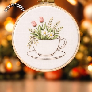 May include: Embroidered floral design in a wooden hoop. The artwork features a white teacup filled with pink tulips, white daisies, and green foliage. The text "ECOCHIC CRAFT" is at the top left of the hoop.