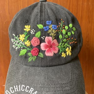 May include: A gray baseball cap with colorful embroidered flowers. The floral design includes pink, yellow, blue, and white blossoms with green leaves. The text "ECOCHICCRAFT" is embroidered on the front of the cap.