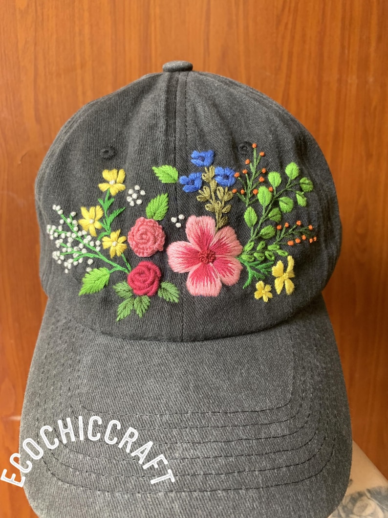 May include: A gray baseball cap with colorful embroidered flowers. The floral design includes pink, yellow, blue, and white blossoms with green leaves. The text "ECOCHICCRAFT" is embroidered on the front of the cap.
