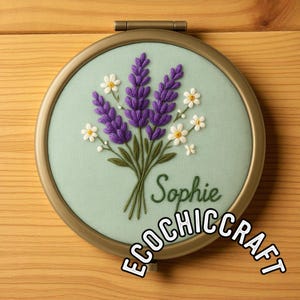 Puede incluir: Arte de aro bordado con un ramo de lavanda morada y margaritas blancas sobre un fondo verde claro. El nombre "Sophie" está bordado en escritura verde. El aro está enmarcado en metal dorado. El texto "ECOCHICCRAFT" también es visible.