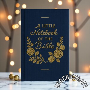 Op de afbeelding: Een marineblauw notitieboek met gouden letters en bloemaccenten. De kaft luidt: "A Little Notebook of the Bible." Het notitieboek staat rechtop, met een vage achtergrond van lichtjes en gouden ornamenten.