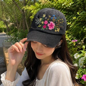 May include: Black baseball cap with embroidered floral design. The cap features a curved brim and a colorful floral arrangement on the front, including pink, yellow, and blue flowers. The cap is made of a soft fabric.