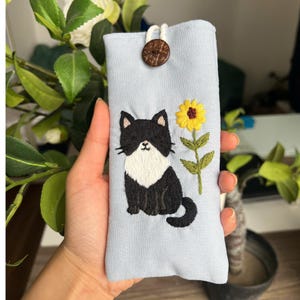 May include: A light blue fabric pouch featuring an embroidered black and white cat sitting next to a yellow sunflower. The pouch is secured at the top with a single brown wooden button and a white cord loop closure.