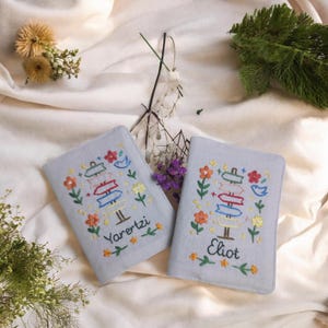 Puede incluir: Dos fundas de pasaporte azul claro con dise&ntilde;os bordados. Cada funda presenta un dise&ntilde;o de poste indicador colorido con flores y los nombres "Yareztzi" y "Eliot". Las fundas est&aacute;n sobre un fondo de tela blanca con detalles florales.