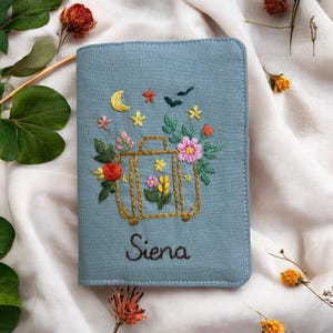 Puede incluir: Un portapasaporte azul claro con un dise&ntilde;o bordado. El dise&ntilde;o presenta una maleta dorada llena de flores coloridas, estrellas, una luna creciente y el nombre "Siena" en escritura.