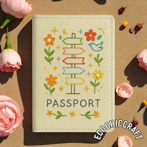 Puede incluir: Una funda de pasaporte beige claro con dise&ntilde;os bordados de flores y se&ntilde;ales direccionales. La funda presenta la palabra "PASSPORT" en una fuente gris oscuro. El dise&ntilde;o incluye elementos naranjas, rojos, amarillos y azules.