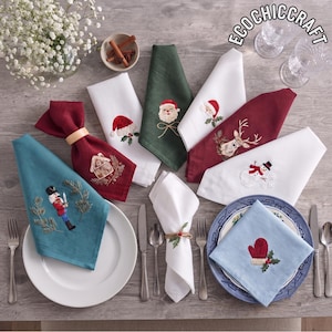May include: A collection of cloth napkins featuring embroidered holiday designs including a nutcracker, Santa Claus, a gingerbread house, a reindeer, a snowman, and a red mitten. The napkins are in shades of teal, white, green, and burgundy.