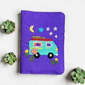 Puede incluir: Funda de pasaporte bordada en morado con un dise&ntilde;o de furgoneta camper turquesa, techo rojo y ruedas negras. El dise&ntilde;o incluye flores, estrellas, una luna y un p&aacute;jaro. La funda est&aacute; rodeada de peque&ntilde;as plantas suculentas.