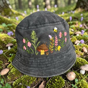 May include: A charcoal gray bucket hat featuring detailed embroidery of a brown mushroom, a small yellow mushroom, pink wildflowers, green foliage, and a small yellow and black bee. The hat has a wide, downward-sloping brim and stitched detailing.