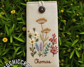 May include: Beige phone case with embroidered floral design. Features a button closure and the name "Chomas" embroidered in brown. The case is decorated with white daisies, red berries, and other colorful flowers. The logo "ECOCHIC CRAFT" is visible.