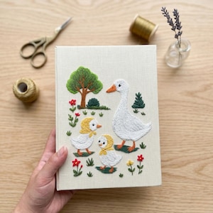 Puede incluir: Un libro rectangular con cubierta de tela color crema que presenta una escena bordada de una oca blanca grande y dos gansitos con capotas amarillas, rodeados por un &aacute;rbol verde, arbustos peque&ntilde;os y flores coloridas rojas y amarillas.