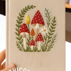 May include: Beige journal with an embroidered design of red mushrooms with white spots, yellow and orange mushrooms, green leaves, and white flowers. The journal is held in a hand.