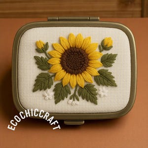 May include: A small, white, rectangular box with a gold metal frame. The box is decorated with an embroidered sunflower with green leaves and white flowers. The text "Ecochiccraft" is printed on the bottom of the box.