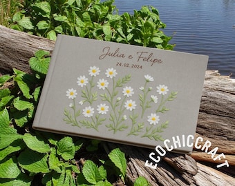 May include: A gray linen guest book with white daisy embroidery and the text "Julia e Felipe 24.02.2024" in brown lettering. The book is on a wooden surface with green foliage.