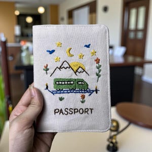 Puede incluir: Funda de pasaporte color crema con dise&ntilde;o bordado. Presenta un tren verde, monta&ntilde;as amarillas, luna y estrellas. La palabra "PASSPORT" est&aacute; bordada en marr&oacute;n. Un accesorio de viaje decorativo.