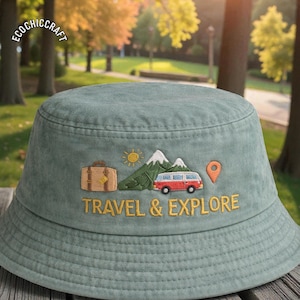 May include: A sage green bucket hat with embroidered travel-themed graphics. The design includes a suitcase, mountains, a red and white van, a sun, and the words "TRAVEL & EXPLORE" in gold thread. The hat is made of a soft, textured fabric.
