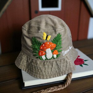 May include: A tan bucket hat featuring an embroidered design of three orange mushrooms with white spots, green fern leaves, and a yellow butterfly. The name Senna is embroidered in white cursive script on the side of the hat crown.